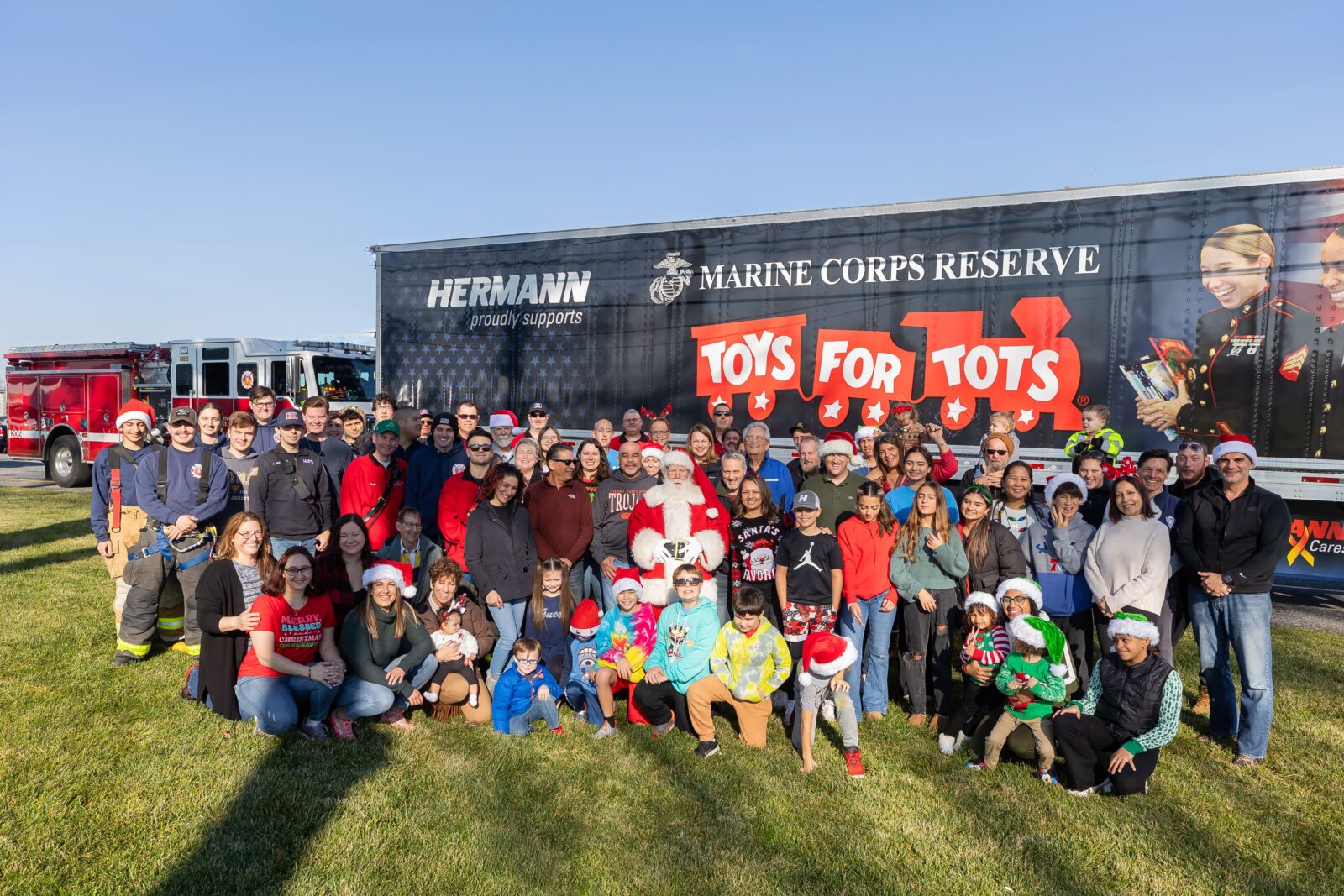 a crowd of happy people stand in front of a trailer with the Toys for Tots logo on it